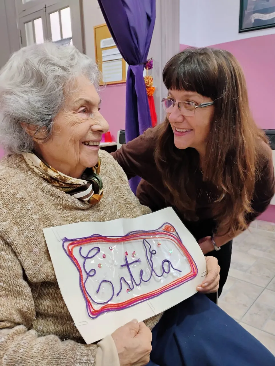 Una concurrente sonríe mostrando un cartel con su nombre, Estela, hecho con lanas de colores en el taller de arte